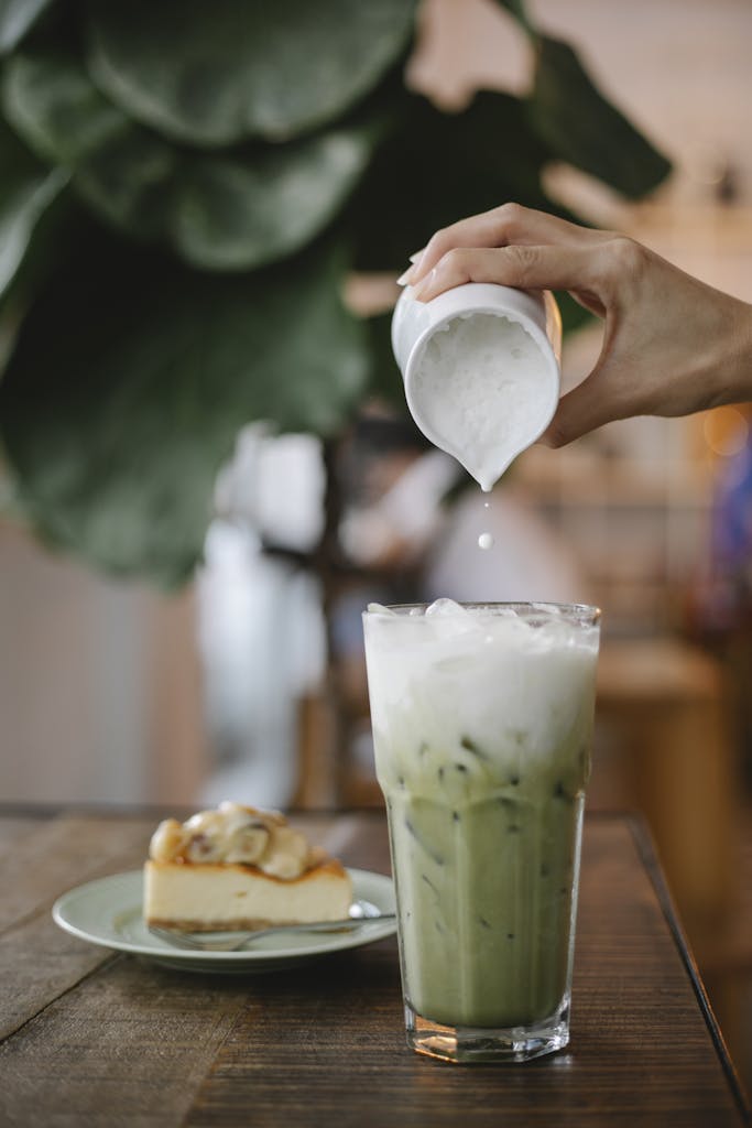 Crop anonymous female pouring whipped milk into glass with matcha frappe placed on table near plate with dessert in cafe